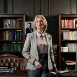 Woman in a blazer standing confidently in an office with bookshelves.